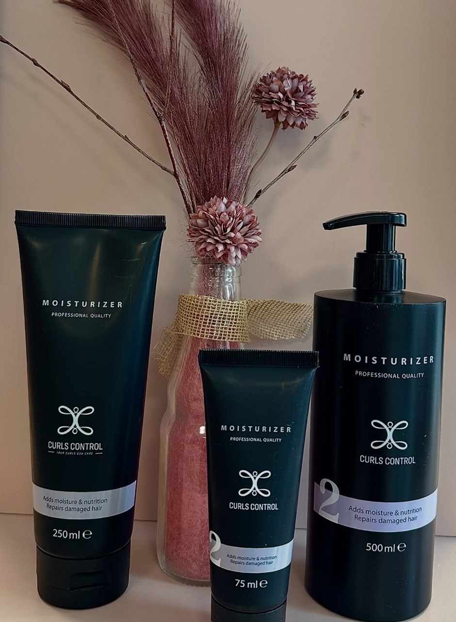 Drie tubes en flessen Curls Control Moisturizer naast een vaas met roze pluimen