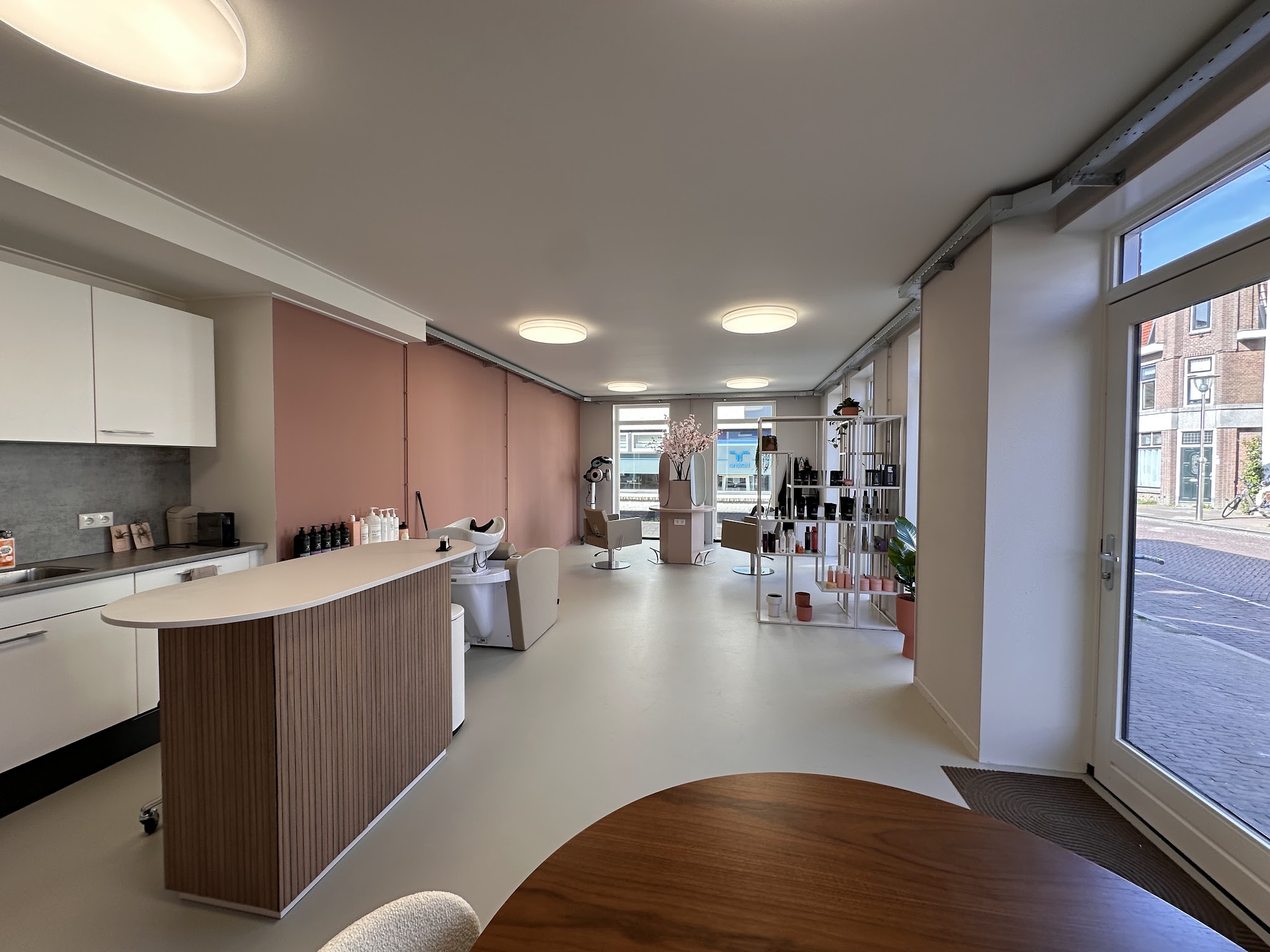 De salon van binnen: een toonbank met houten latten, de roze wand en grote ramen aan de straat